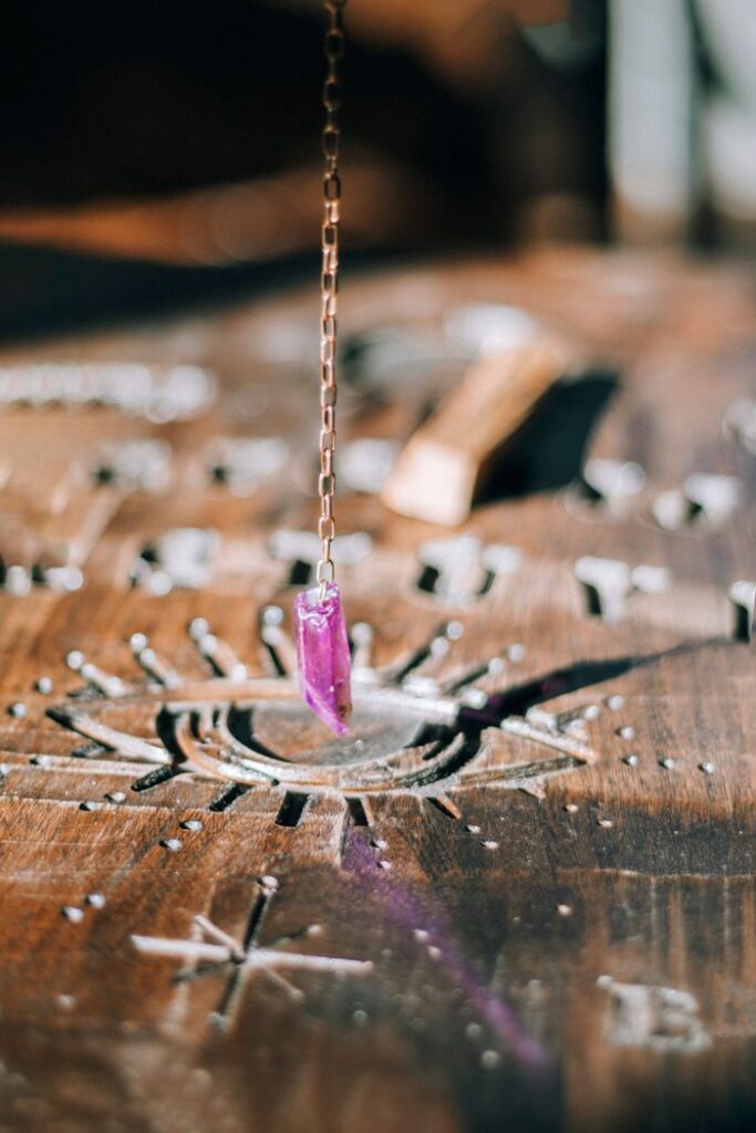 A close-up of a purple crystal pendant hanging on a chain above a carved wooden board, possibly for divination or spiritual practices.